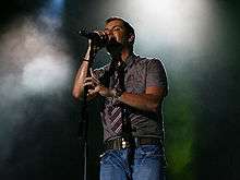 A man holding a microphone pedestal, wearing a coffee-colored shirt and jeans.
