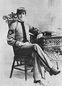 A ornate chair and a table with a book on it. A man sits in the chair, wearing an American Civil War style peaked cap. On his sleeves he wears three stripes pointed down with a lozenge of a First Sergeant.