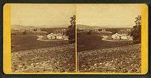Two nearly identical images side by side in stereograph format of a small 19th century college campus.  In the foreground are neatly planted rows of crops.  The nearest building is an ornate 19th century conservatory or greenhouse.  In the distance, on a ridge, are three brick academic buildings.