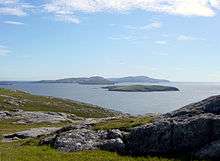 A rocky moorland in the foreground and low, dark islands against a blue sky in the background.