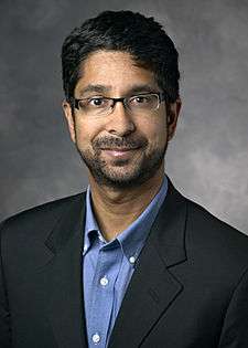 A portrait of Vijay Pande, looking straight ahead. His ethnicity is Indian. He has medium-length black hair, black-rim glasses, and a short mustache and beard. He is wearing a blue polo shirt under a black suit coat.