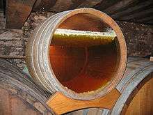 Photo showing a barrel with a glass bottom revealing the layer of yeast on the surface under which the yellow wine is ageing. This yeast is in the form of an irregular white layer - it forms small stalactites that sink a few millimetres into the wine which is already dark yellow wine.  The wall in the background is grey, probably coated in grey mould.