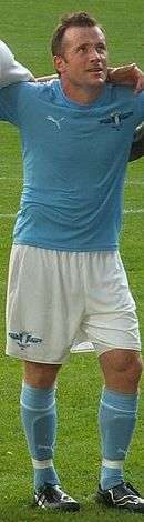 A dark haired man stands with his arms around the shoulders of two team-mates, each of whom is out of shot. The subject wears a light blue shirt, white shorts and light blue socks.