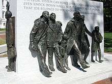 Bronze sculptures of seven figures marching stand around a large rectangular block of white engraved granite.