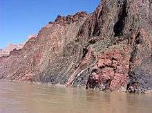 Gray and reddish rock face with rough surface adjacent to a river.