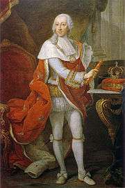 Full-length portrait of a bewigged monarch in white tights and a red robe with his crown sitting on a table