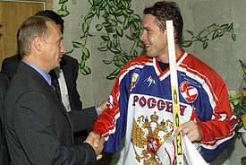 An ice hockey player in his early thirties shakes hands with a middle-aged man dressed in a dark suit. The hockey player wears a white, red and blue jersey labeled "РОССИЯ" and holds a hockey stick.