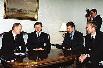 Four men in suits, two middle-aged and two in their thirties, sit smiling around a glossy wood table. A photographer hovers in the background.