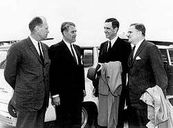 Four men in suits are outdoors, speaking to each other in front of a large white automobile.