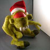 A green plush toy of a Vortigaunt sits against a gray wall. The toy is wearing a Santa Claus hat, leaving only the dominant red eye visible