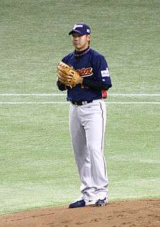 A young Japanese man wearing a dark blue and grey Japan national baseball uniform stands on a pitching mound.