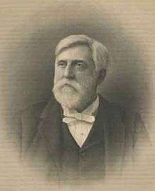 A white-haired man with a beard and mustache, facing left. He is wearing a white shirt, black vest and black jacket