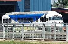 A single train car, painted blue and white, sits on a track between a metal fence and a large blue building.