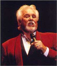 A man with a gray mustache, beard, and hair, wearing a red jacket and singing into a microphone