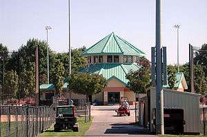 A hexagonal building with a green roof is partly surrounded by trees, fences, and lights on tall poles. A man in a baseball-style cap is driving a small machine across a paved surface near the hexagonal building. Another machine with the letters T-O-R-O on its rear bumper is parked on the grass nearby.