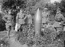 A group photograph of several men in uniform who are standing round an unexploded shell that is standing pointed-end up from the ground.