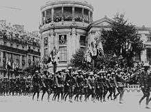black & white photograph of Marines in a formation marching through a French street with French buildings in the background, decorated with the flags of allied nations