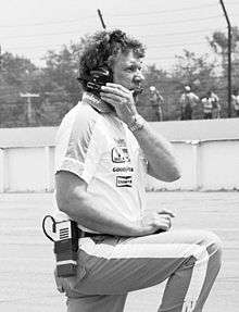 man standing on racetrack pit lane talking to his driver on a headset