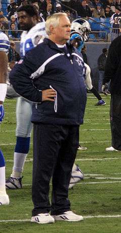 Candid photograph of Phillips standing with arms akimbo on a football field wearing a dark blue jacket with a Dallas Cowboys logo