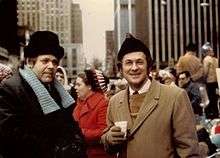 Two men in winter coats and hats standing in crowd in front of tall city buildings