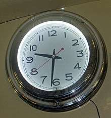 A shiny metallic circular clock with a face illuminated from within, marked in plain numerals in a sans-serif font, showing 9:32