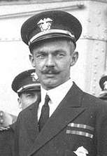 Head and shoulders of a white man with a handlebar mustache, wearing a peaked cap and a dark jacket with two rows of ribbon bars on the left breast. Several other men in similar clothing can be seen behind him.