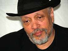 Head and shoulders of man with drooping eyelids wearing black fedora, black shirt without a collar, black jacket, and mostly grey short trimmed beard.