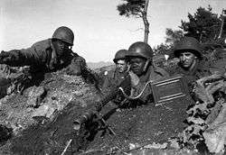 Four soldiers sitting in a foxhole, one soldier is pointing a machine gun towards the camera while another is pointing out targets