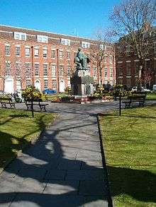 Statue on a plinth in park area, with buildings behind.