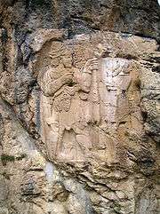 The photograph shows a sculpted rock: a bas-relief appears in grey-beige on a blue-gray rock stained with dark grey streaks.  The Hittite king is on the left.  He is bearded and holding a branch with two grapes.  Facing him, the god has lost his head and his feet because of the ravages of time
