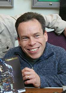 Actor Warwick Davis sits at a table, smiling and holding Leprechaun merchandise
