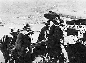 A group of men in slouch hats struggle with a large object. In the background is a jeep and a Dakota.