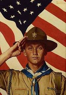 A Boy Scout in full uniform stands saluting while an American Flag waves in the background.