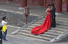 A woman wearing a long, ornate red dress stands next to a man in a black suit on some short stone steps while another man photographs them from the foot of the stairs