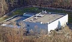 A large light-colored building with a flat roof seen from above, surrounded by bare trees.