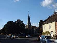 Street scene with a church and spire central to the picture. To the right is a yellow building with a pub sign. To the left is a large tree with a signpost in front. Several cars.