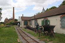 Single storey buildings, with a parallel railway track in front. Between them is a ramp. A chimney can be seen in the distance.