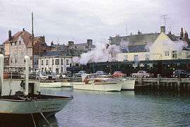 A green pannier tank locomotive is pulling green passenger coaches past a row of parked cars. In the foreground is a harbour with moored pleasure craft. Behind the train is a row of buildings.