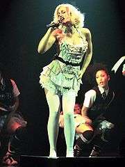 A blonde lady in a pale green dress with black belt, white tights and white shoes sings into a microphone. In the background is a Japanese dancer wearing a school uniform-style outfit: white shirt, black waitcoat, tie and red-and-black tartan skirt with white mesh underneath. The dancer is also wearing black sport socks and shoes. To the left of the image, parts of another dancer is visible; she is dressed similarly to the first dancer.
