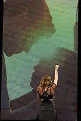 A female teenager with long brown hair and a black dress stretches her fist upward. Only the teen's back is visible, as she is facing a large screen behind her. On the screen is the silhouette of a male teen caressing the face of a shorter female teenager.