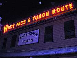 A red electric sign is seen at night, displaying words "White Pass & Yukon Route" above a white, wooden sign emblazoned with "Whitehorse Yukon".
