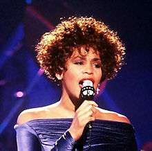 A black woman short curly brown hair wearing a purple dress sings to a microphone