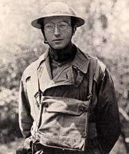 Head and torso of a man wearing a metal helmet with wide brim, wire frame glasses, a military coat, and a satchel loosely strapped to his chest.