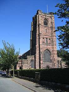 The west end of a Gothic Revival style church built in different coloured sandstone, showing a tall tower with a flagpole; in front is a hedge and to the sides are trees