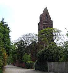 A tall narrow tower with a small pointed spire surrounded by trees in leaf almost hiding the body of the church; to the left is a footpath with railings and a wall