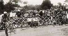 A group of around ten cyclists, climbing over a fence. Behind the fence are cars.