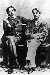 Two men sit on a bench with their legs crossed. Both are well dressed in suits.