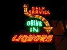 A green and orange neon sign that says "self service drive in liquors," and has an arrow pointing downward.