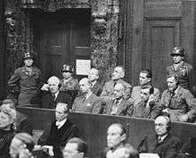 a black and white photograph of a US court room in Nuremberg with several rows of German defendants sitting on a tiered stand with guards standing behind them