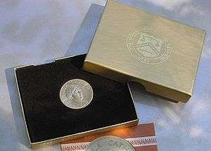 A gold medallion in a box, including a lid and a piece of paper